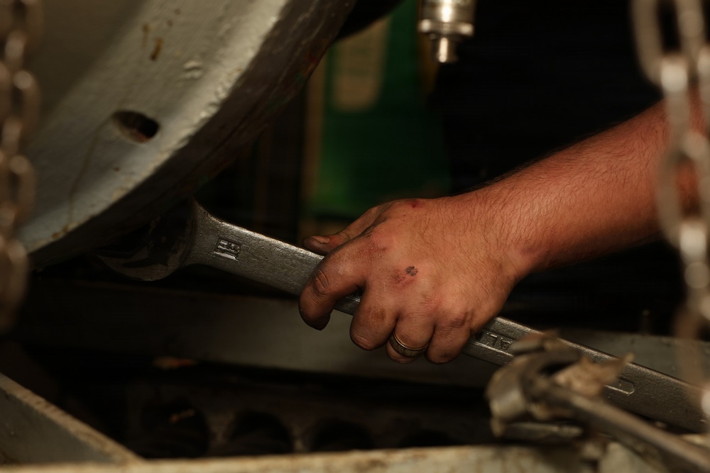 A close-up photograph of a mechanic's hand gripping a large open-end wrench while working on machinery or a vehicle engine. The hand shows signs of manual labor including dirt, minor cuts, and a ring on one finger. The setting appears to be an automotive repair shop or garage.