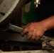 A close-up photograph of a mechanic's hand gripping a large open-end wrench while working on machinery or a vehicle engine. The hand shows signs of manual labor including dirt, minor cuts, and a ring on one finger. The setting appears to be an automotive repair shop or garage.