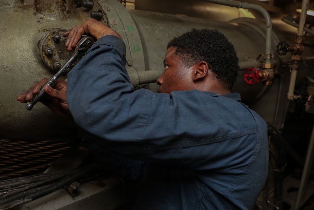 A technician or mechanic in a blue coverall uniform is using a ratchet wrench to work on a large cylindrical piece of industrial or naval machinery. The setting appears to be an engine room or mechanical space, likely aboard a ship, indicated by the dense piping, valves, and cable bundles visible in the background. The worker is focused intently on tightening or loosening bolts on a flanged connection.
