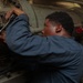 A technician or mechanic in a blue coverall uniform is using a ratchet wrench to work on a large cylindrical piece of industrial or naval machinery. The setting appears to be an engine room or mechanical space, likely aboard a ship, indicated by the dense piping, valves, and cable bundles visible in the background. The worker is focused intently on tightening or loosening bolts on a flanged connection.
