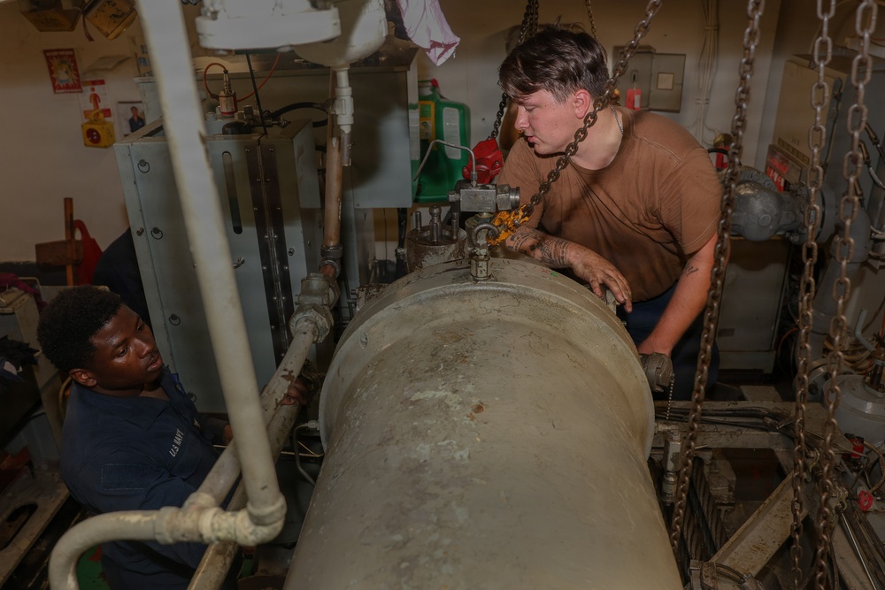 Two U.S. Navy sailors are working on a large cylindrical piece of equipment in what appears to be a ship's machinery or engineering space. One sailor in a brown t-shirt is leaning over the cylinder manipulating a chain hoist or lifting mechanism, while a second sailor in a Navy blue uniform observes from the left side. The workspace is cluttered with pipes, chains, electrical panels, and various maintenance equipment.