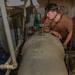 Two U.S. Navy sailors are working on a large cylindrical piece of equipment in what appears to be a ship's machinery or engineering space. One sailor in a brown t-shirt is leaning over the cylinder manipulating a chain hoist or lifting mechanism, while a second sailor in a Navy blue uniform observes from the left side. The workspace is cluttered with pipes, chains, electrical panels, and various maintenance equipment.