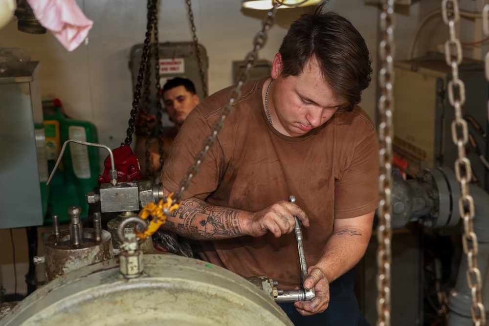 A young male mechanic with tattoos on his forearms is using a ratchet wrench to work on heavy industrial equipment in what appears to be a ship's engine room or mechanical workspace. He is wearing a brown t-shirt soiled with grease and is focused on the task at hand. A second worker is visible in the background observing or assisting.