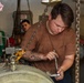A young male mechanic with tattoos on his forearms is using a ratchet wrench to work on heavy industrial equipment in what appears to be a ship's engine room or mechanical workspace. He is wearing a brown t-shirt soiled with grease and is focused on the task at hand. A second worker is visible in the background observing or assisting.