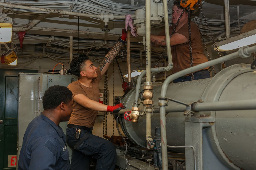Three military personnel, likely U.S. Navy sailors, are performing maintenance work in what appears to be an engine room or mechanical space aboard a naval vessel. Two individuals wearing brown t-shirts are working on overhead piping and equipment, while a third in a dark uniform observes. The space is densely packed with pipes, cables, valves, and mechanical components typical of a ship's engineering spaces.