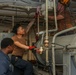 Three military personnel, likely U.S. Navy sailors, are performing maintenance work in what appears to be an engine room or mechanical space aboard a naval vessel. Two individuals wearing brown t-shirts are working on overhead piping and equipment, while a third in a dark uniform observes. The space is densely packed with pipes, cables, valves, and mechanical components typical of a ship's engineering spaces.
