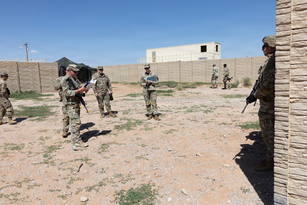 A group of military personnel in camouflage uniforms are gathered in an open compound area, appearing to conduct a briefing or after-action review. Several soldiers are holding documents or clipboards while others stand at the perimeter. The setting appears to be a training facility or forward operating base with concrete walls and temporary structures.