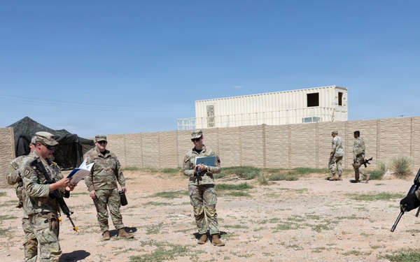 A group of military personnel in camouflage uniforms are gathered in an open compound area, appearing to conduct a briefing or after-action review. Several soldiers are holding documents or clipboards while others stand at the perimeter. The setting appears to be a training facility or forward operating base with concrete walls and temporary structures.