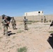A group of military personnel in camouflage uniforms are gathered in an open compound area, appearing to conduct a briefing or after-action review. Several soldiers are holding documents or clipboards while others stand at the perimeter. The setting appears to be a training facility or forward operating base with concrete walls and temporary structures.