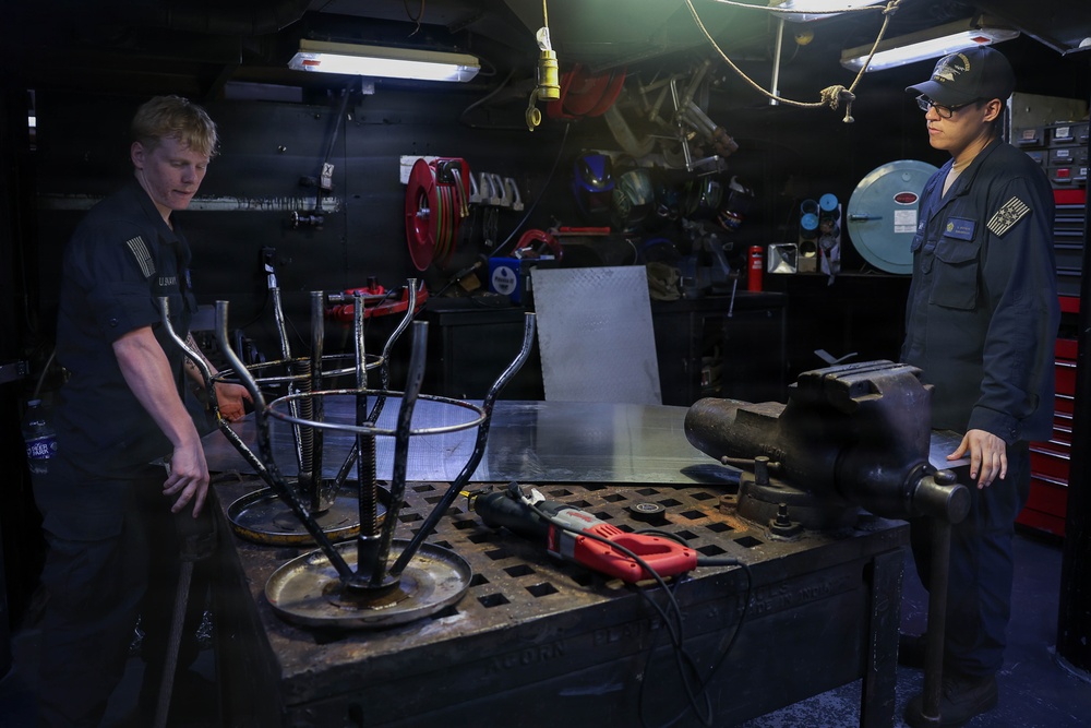 Two individuals in dark military-style uniforms are working in what appears to be a shipboard or industrial workshop/machine shop. They are examining a metal fabrication piece on a heavy-duty work table. The workspace is equipped with various tools, equipment, and storage units typical of a maintenance or fabrication facility.