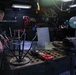 Two individuals in dark military-style uniforms are working in what appears to be a shipboard or industrial workshop/machine shop. They are examining a metal fabrication piece on a heavy-duty work table. The workspace is equipped with various tools, equipment, and storage units typical of a maintenance or fabrication facility.