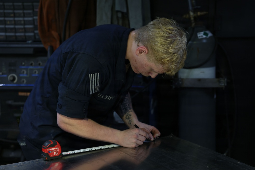 A U.S. Navy service member in dark blue coveralls is bent over a metal work surface, marking measurements with a marker. A Craftsman tape measure (16-foot) is visible on the surface beside them. The setting appears to be a shipboard or industrial workshop environment.