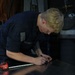 A U.S. Navy service member in dark blue coveralls is bent over a metal work surface, marking measurements with a marker. A Craftsman tape measure (16-foot) is visible on the surface beside them. The setting appears to be a shipboard or industrial workshop environment.