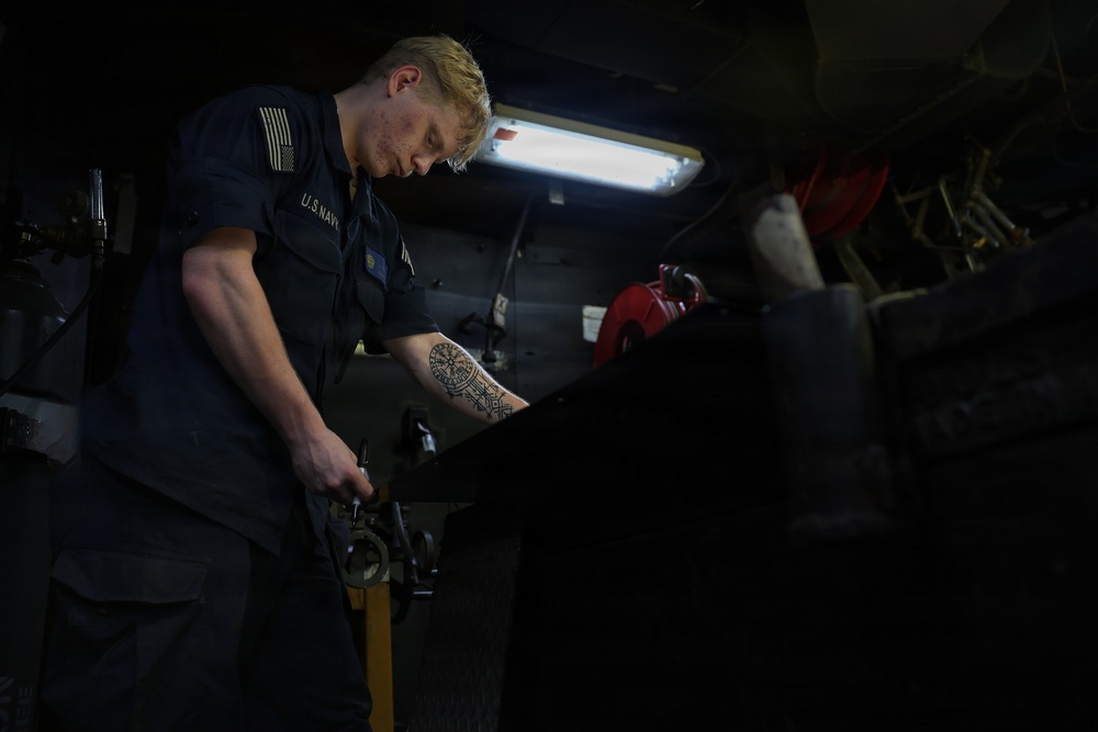 A U.S. Navy sailor is working in what appears to be a confined mechanical or engineering space aboard a naval vessel. The individual is focused on a task involving a hand tool, working over a dark surface in a dimly lit compartment. A detailed geometric tattoo is visible on their forearm.