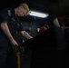 A U.S. Navy sailor is working in what appears to be a confined mechanical or engineering space aboard a naval vessel. The individual is focused on a task involving a hand tool, working over a dark surface in a dimly lit compartment. A detailed geometric tattoo is visible on their forearm.