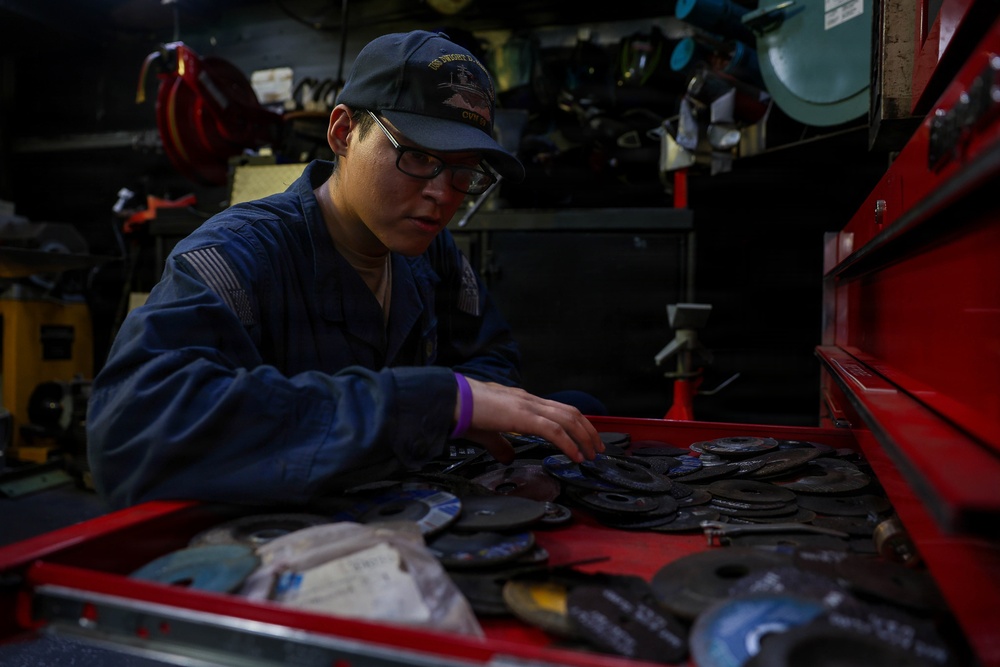 A person wearing a dark blue military-style uniform and a navy cap is working in what appears to be a shipboard or industrial workshop. They are sorting through a red tool chest drawer filled with numerous grinding/cutting discs. The workspace is cluttered with various tools and equipment visible in the background.