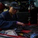 A person wearing a dark blue military-style uniform and a navy cap is working in what appears to be a shipboard or industrial workshop. They are sorting through a red tool chest drawer filled with numerous grinding/cutting discs. The workspace is cluttered with various tools and equipment visible in the background.