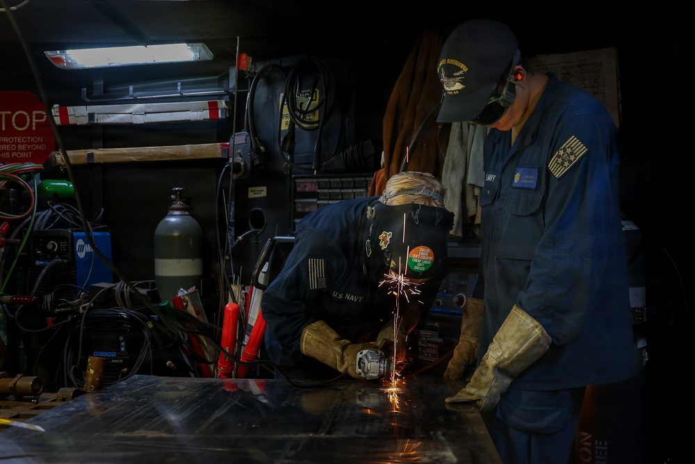 Two U.S. Navy personnel are working in what appears to be a shipboard metalworking or welding shop. One sailor is actively grinding or cutting metal on a work surface, producing visible sparks, while the other observes and supervises. Both are wearing blue Navy coveralls and protective equipment in a cluttered industrial workspace.