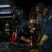 Two U.S. Navy personnel are working in what appears to be a shipboard metalworking or welding shop. One sailor is actively grinding or cutting metal on a work surface, producing visible sparks, while the other observes and supervises. Both are wearing blue Navy coveralls and protective equipment in a cluttered industrial workspace.