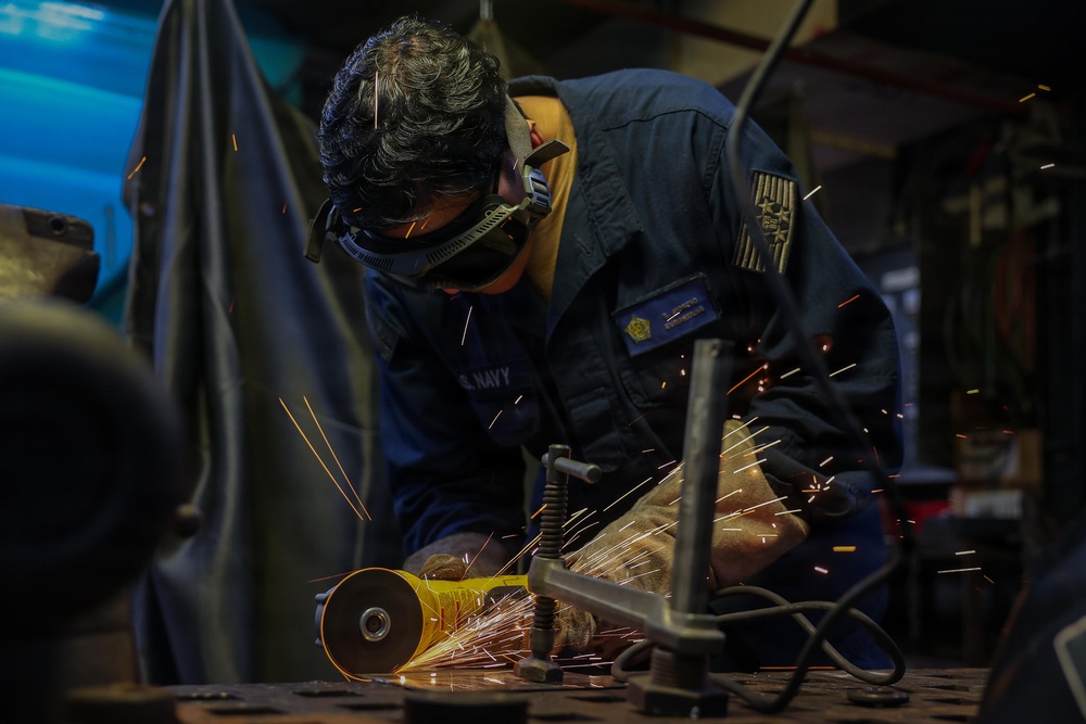 A U.S. Navy sailor in blue coveralls is using an angle grinder on a metal workpiece, producing a shower of bright sparks. The worker is wearing protective goggles and gloves in what appears to be a shipboard or industrial workshop environment. The image captures the intensity of the metalworking task with dramatic lighting from the sparks.