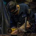 A U.S. Navy sailor in blue coveralls is using an angle grinder on a metal workpiece, producing a shower of bright sparks. The worker is wearing protective goggles and gloves in what appears to be a shipboard or industrial workshop environment. The image captures the intensity of the metalworking task with dramatic lighting from the sparks.