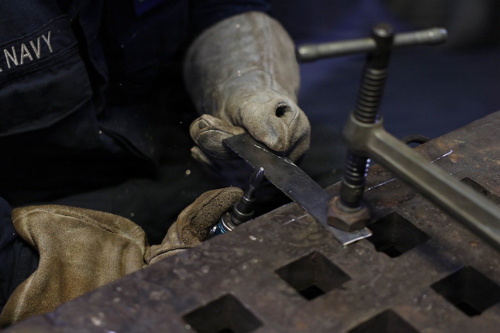 A U.S. Navy personnel member is performing metalwork on a welding or fabrication table, using a drill or rotary tool on a metal piece secured by a C-clamp. The worker is wearing worn leather protective gloves and a dark Navy-issued uniform. The work surface appears to be a heavy-duty steel grating table commonly found in a shipboard or industrial workshop environment.