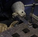A U.S. Navy personnel member is performing metalwork on a welding or fabrication table, using a drill or rotary tool on a metal piece secured by a C-clamp. The worker is wearing worn leather protective gloves and a dark Navy-issued uniform. The work surface appears to be a heavy-duty steel grating table commonly found in a shipboard or industrial workshop environment.