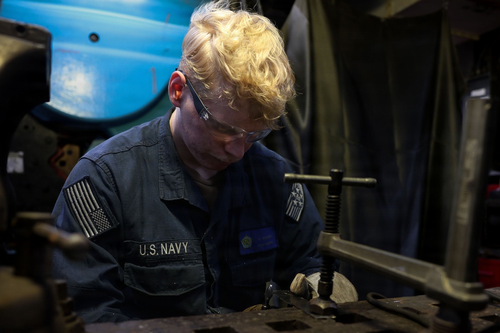A U.S. Navy sailor with blonde curly hair is focused on mechanical work at a workbench in what appears to be a ship's machine shop or maintenance space. The sailor is wearing a dark blue Navy coverall uniform with a U.S. flag patch and 'U.S. NAVY' name tape, along with clear safety glasses. The individual appears to be working with a vise or clamp on a metal component.