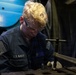 A U.S. Navy sailor with blonde curly hair is focused on mechanical work at a workbench in what appears to be a ship's machine shop or maintenance space. The sailor is wearing a dark blue Navy coverall uniform with a U.S. flag patch and 'U.S. NAVY' name tape, along with clear safety glasses. The individual appears to be working with a vise or clamp on a metal component.