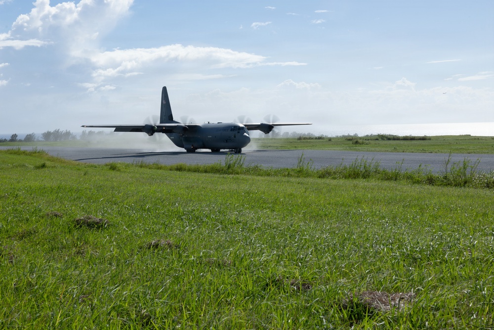 A military turboprop transport aircraft, consistent with a C-130 Hercules design, is seen on a runway either landing or taxiing, with propeller wash kicking up dust and debris behind it. The aircraft is painted in a dark gray military color scheme. The scene is set at a relatively simple airstrip surrounded by green grass and trees under a partly cloudy sky.