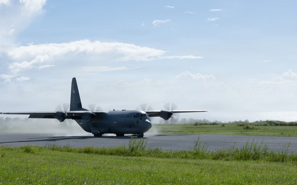 A military turboprop transport aircraft, consistent with a C-130 Hercules design, is seen on a runway either landing or taxiing, with propeller wash kicking up dust and debris behind it. The aircraft is painted in a dark gray military color scheme. The scene is set at a relatively simple airstrip surrounded by green grass and trees under a partly cloudy sky.