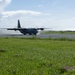A military turboprop transport aircraft, consistent with a C-130 Hercules design, is seen on a runway either landing or taxiing, with propeller wash kicking up dust and debris behind it. The aircraft is painted in a dark gray military color scheme. The scene is set at a relatively simple airstrip surrounded by green grass and trees under a partly cloudy sky.