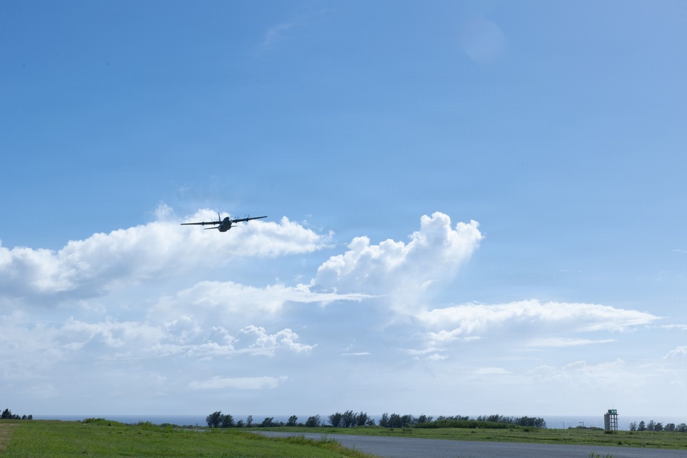 A turboprop aircraft, likely a military transport such as a C-130 Hercules, is captured in flight at low altitude above what appears to be a small airfield or runway. The scene is set against a bright blue sky with scattered cumulus clouds, with a tree line and a small observation or control tower visible on the horizon. The aircraft appears to be either taking off or performing a low-altitude pass.