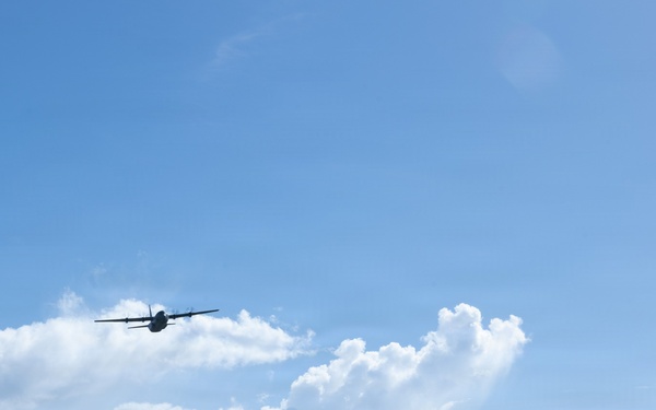 A turboprop aircraft, likely a military transport such as a C-130 Hercules, is captured in flight at low altitude above what appears to be a small airfield or runway. The scene is set against a bright blue sky with scattered cumulus clouds, with a tree line and a small observation or control tower visible on the horizon. The aircraft appears to be either taking off or performing a low-altitude pass.