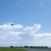A turboprop aircraft, likely a military transport such as a C-130 Hercules, is captured in flight at low altitude above what appears to be a small airfield or runway. The scene is set against a bright blue sky with scattered cumulus clouds, with a tree line and a small observation or control tower visible on the horizon. The aircraft appears to be either taking off or performing a low-altitude pass.