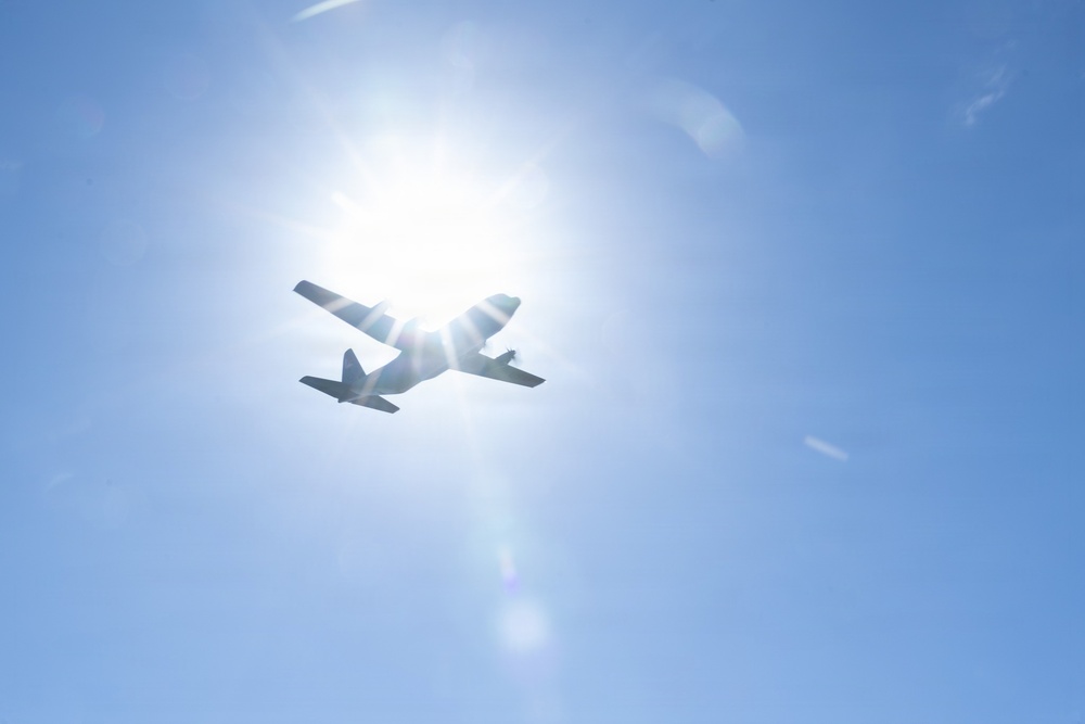 A low-angle photograph captures an aircraft in silhouette flying directly in front of the sun against a clear blue sky. The strong backlight creates a dramatic starburst lens flare effect, rendering the aircraft almost entirely as a dark silhouette. The image appears to be taken from the ground looking upward.