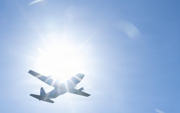 A low-angle photograph captures an aircraft in silhouette flying directly in front of the sun against a clear blue sky. The strong backlight creates a dramatic starburst lens flare effect, rendering the aircraft almost entirely as a dark silhouette. The image appears to be taken from the ground looking upward.