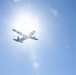 A low-angle photograph captures an aircraft in silhouette flying directly in front of the sun against a clear blue sky. The strong backlight creates a dramatic starburst lens flare effect, rendering the aircraft almost entirely as a dark silhouette. The image appears to be taken from the ground looking upward.