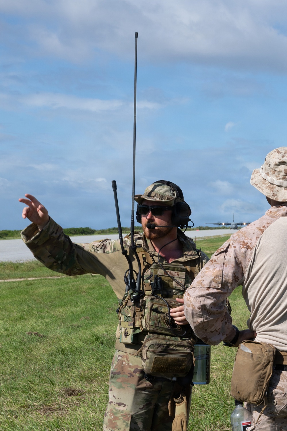 Two military personnel are standing near an airfield runway, engaged in a briefing or coordination discussion. The primary figure, identified by a name tape reading 'MOHLER,' is wearing OCP (Operational Camouflage Pattern) uniform with a tactical vest, communications headset, and multiple radio antennas. A military transport aircraft, consistent with a C-130, is visible on the runway in the background.