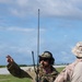 Two military personnel are standing near an airfield runway, engaged in a briefing or coordination discussion. The primary figure, identified by a name tape reading 'MOHLER,' is wearing OCP (Operational Camouflage Pattern) uniform with a tactical vest, communications headset, and multiple radio antennas. A military transport aircraft, consistent with a C-130, is visible on the runway in the background.