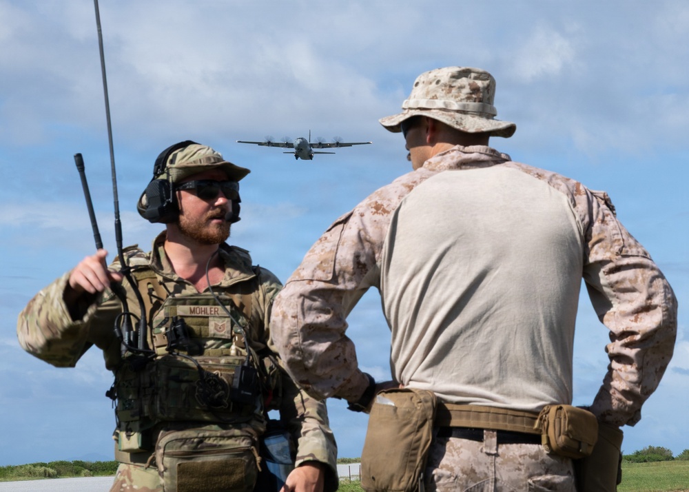 Two military personnel are observed on what appears to be an airfield or open terrain, with a military transport aircraft visible in the background sky. The individual on the left, identified by a name tape as 'MOHLER,' is wearing Air Force multicam tactical gear with a radio antenna and communications headset, while the individual on the right wears USMC MARPAT desert camouflage. The scene suggests a joint military exercise or operation involving air-ground coordination.
