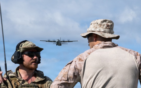 Two military personnel are observed on what appears to be an airfield or open terrain, with a military transport aircraft visible in the background sky. The individual on the left, identified by a name tape as 'MOHLER,' is wearing Air Force multicam tactical gear with a radio antenna and communications headset, while the individual on the right wears USMC MARPAT desert camouflage. The scene suggests a joint military exercise or operation involving air-ground coordination.