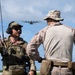 Two military personnel are observed on what appears to be an airfield or open terrain, with a military transport aircraft visible in the background sky. The individual on the left, identified by a name tape as 'MOHLER,' is wearing Air Force multicam tactical gear with a radio antenna and communications headset, while the individual on the right wears USMC MARPAT desert camouflage. The scene suggests a joint military exercise or operation involving air-ground coordination.