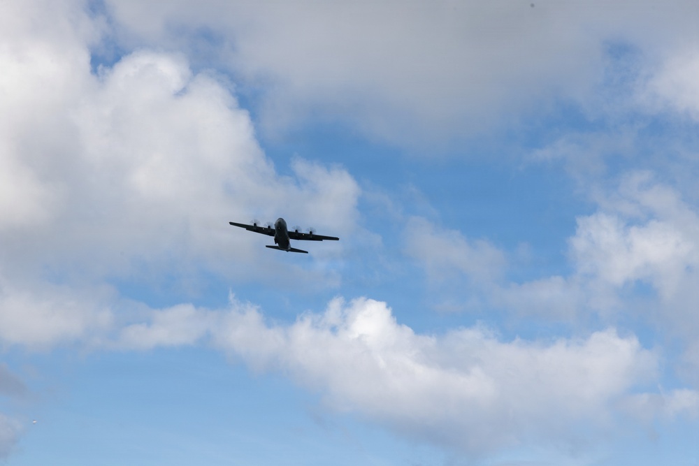 A large multi-engine turboprop military transport aircraft is photographed in flight against a partly cloudy blue sky. The aircraft is viewed from below at a slight angle, appearing as a dark silhouette. The image captures the aircraft in level cruise flight with no visible markings discernible at this distance.