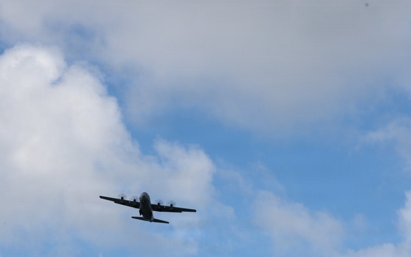 A large multi-engine turboprop military transport aircraft is photographed in flight against a partly cloudy blue sky. The aircraft is viewed from below at a slight angle, appearing as a dark silhouette. The image captures the aircraft in level cruise flight with no visible markings discernible at this distance.