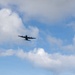 A large multi-engine turboprop military transport aircraft is photographed in flight against a partly cloudy blue sky. The aircraft is viewed from below at a slight angle, appearing as a dark silhouette. The image captures the aircraft in level cruise flight with no visible markings discernible at this distance.