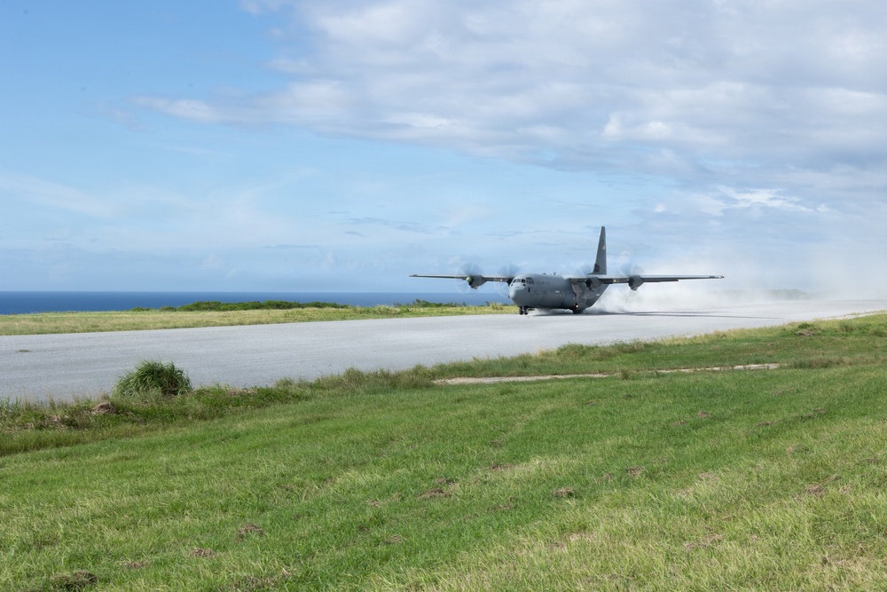 A military turboprop transport aircraft, consistent with a C-130 Hercules, is landing or taking off on a short runway near a coastline. The aircraft is generating significant dust and spray behind it, suggesting high-speed ground contact. The setting appears to be a remote or austere airstrip surrounded by grass and ocean in the background.