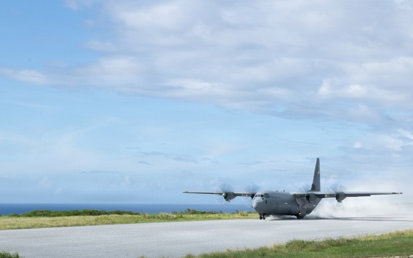 A military turboprop transport aircraft, consistent with a C-130 Hercules, is landing or taking off on a short runway near a coastline. The aircraft is generating significant dust and spray behind it, suggesting high-speed ground contact. The setting appears to be a remote or austere airstrip surrounded by grass and ocean in the background.