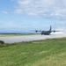 A military turboprop transport aircraft, consistent with a C-130 Hercules, is landing or taking off on a short runway near a coastline. The aircraft is generating significant dust and spray behind it, suggesting high-speed ground contact. The setting appears to be a remote or austere airstrip surrounded by grass and ocean in the background.