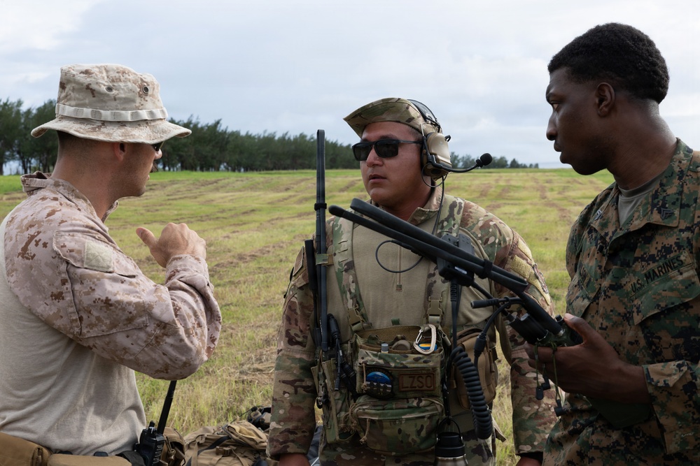 Three military personnel are engaged in a discussion in an open field environment. Two individuals wear U.S. Marine Corps uniforms (desert MARPAT and woodland MARPAT patterns), while the center individual wears Army MultiCam/OCP pattern with tactical communications gear. The setting appears to be a training exercise or operational briefing in a grassy field.